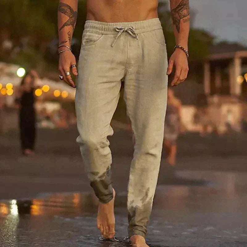 Man wearing beige lightweight linen drawstring trousers with side pockets, standing on a beach at dusk.
