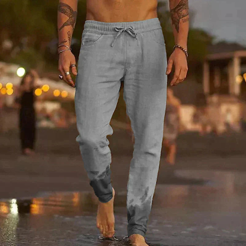 Grey men's lightweight linen trousers with an adjustable drawstring waist, worn barefoot on a beach.