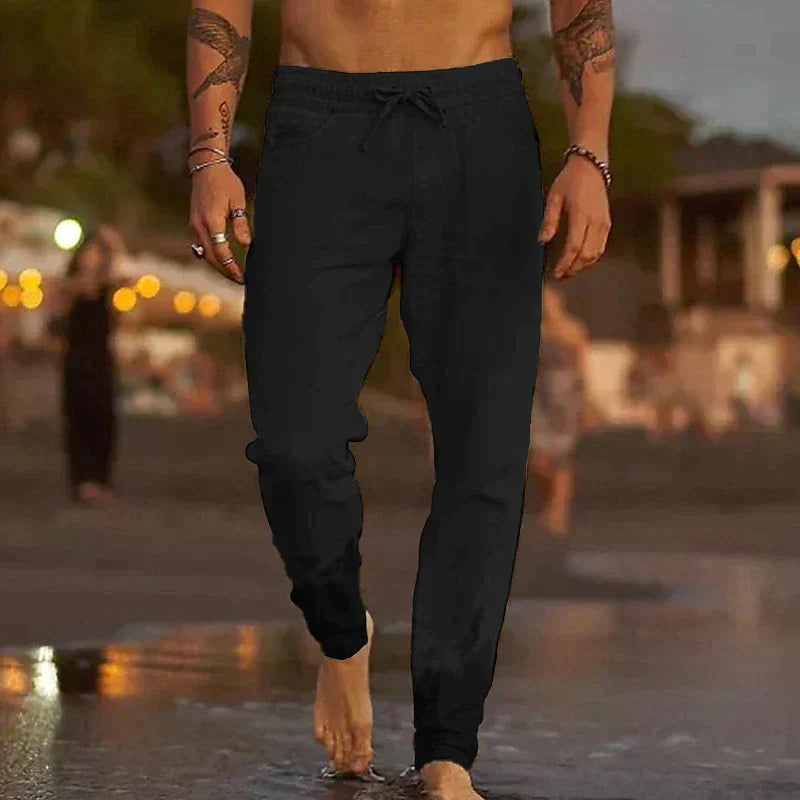 Black lightweight linen trousers with a relaxed fit, drawstring waist, and side pockets worn on a beach.