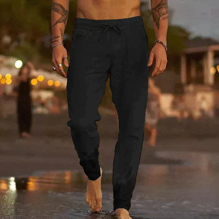 Black lightweight linen trousers with a relaxed fit, drawstring waist, and side pockets worn on a beach.