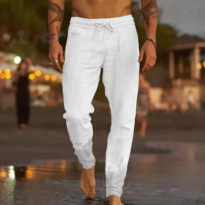 White lightweight linen drawstring trousers for men with a relaxed fit, worn barefoot on a beach.