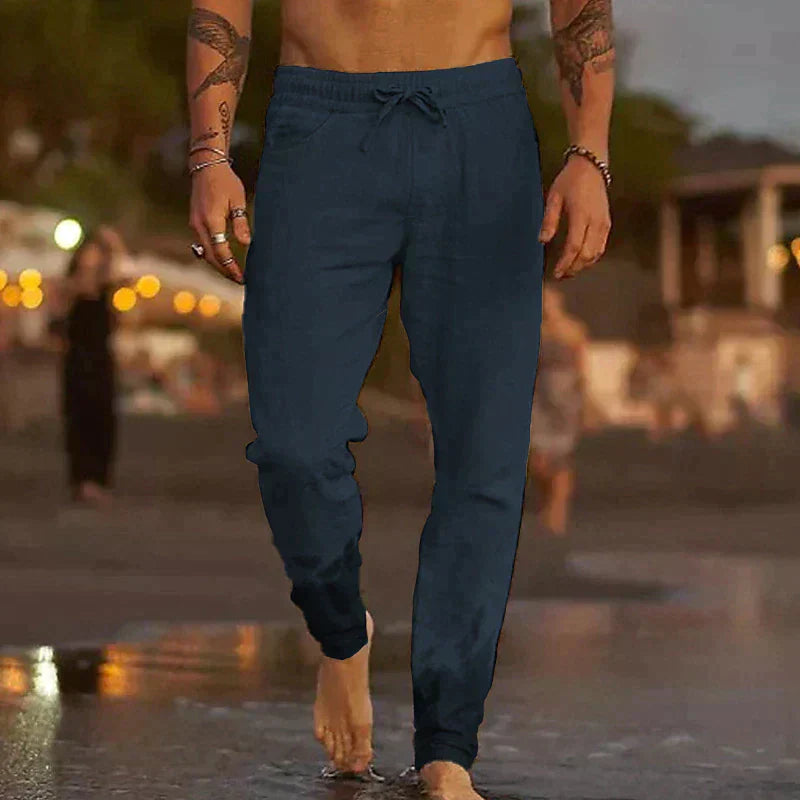 Men’s navy blue linen drawstring trousers with a relaxed fit, worn barefoot on a beach at twilight.