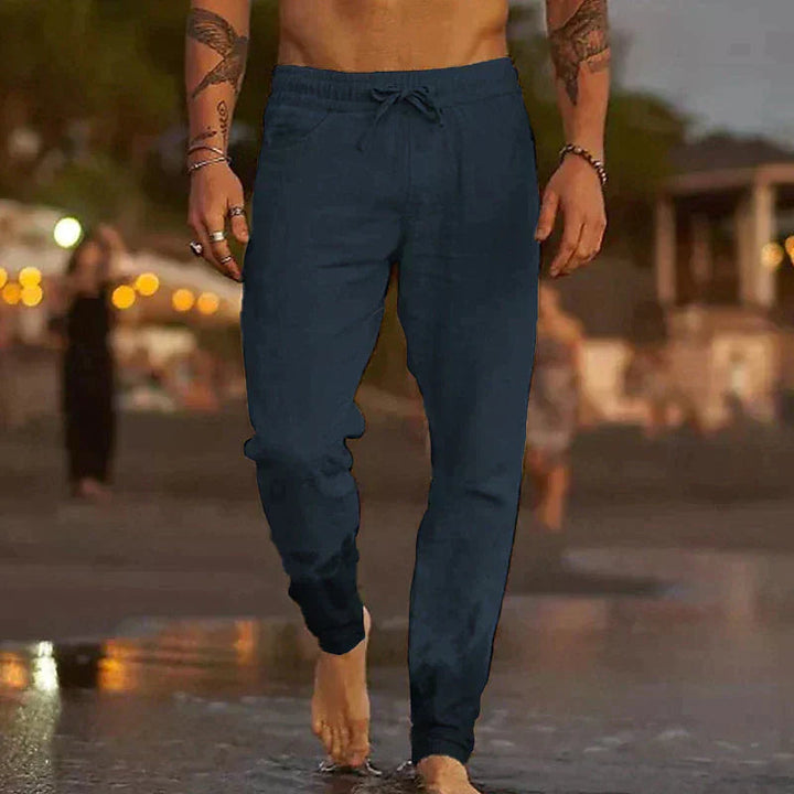 Men’s navy blue linen drawstring trousers with a relaxed fit, worn barefoot on a beach at twilight.