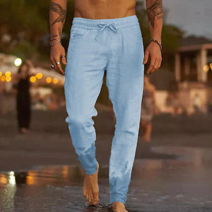Light blue men's linen trousers with a relaxed fit, side pockets, and an adjustable drawstring waist on a beach.