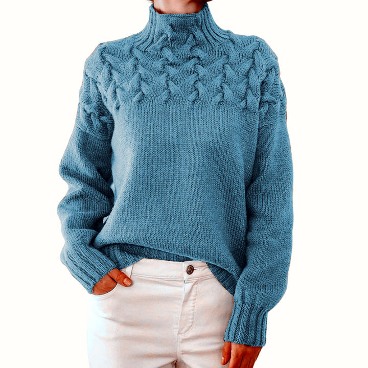 Blue cable knit turtleneck jumper with a braided yoke design, relaxed fit, and ribbed cuffs, worn with white trousers.