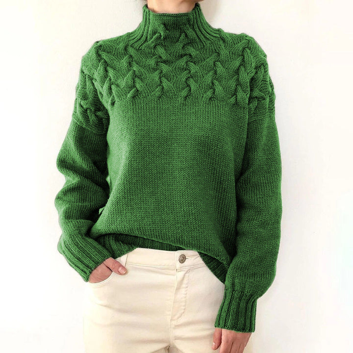 Green cable knit turtleneck jumper with a braided yoke, relaxed fit, and ribbed cuffs worn with cream trousers.