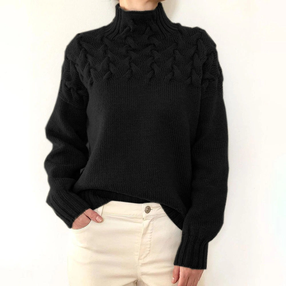 Black cable knit turtleneck jumper with a braided yoke, relaxed fit, and ribbed cuffs worn with cream trousers.