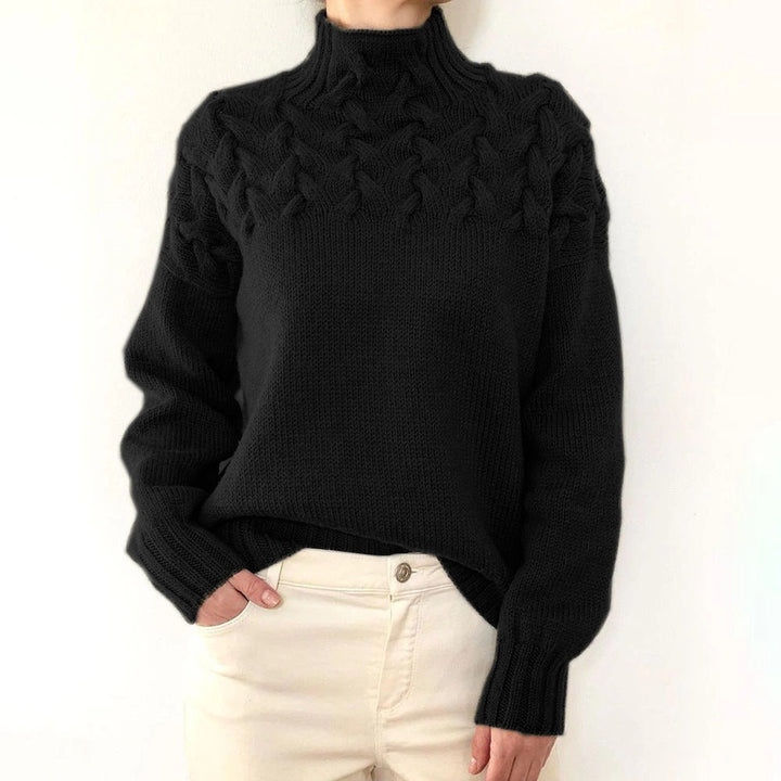 Black cable knit turtleneck jumper with a braided yoke, relaxed fit, and ribbed cuffs worn with cream trousers.