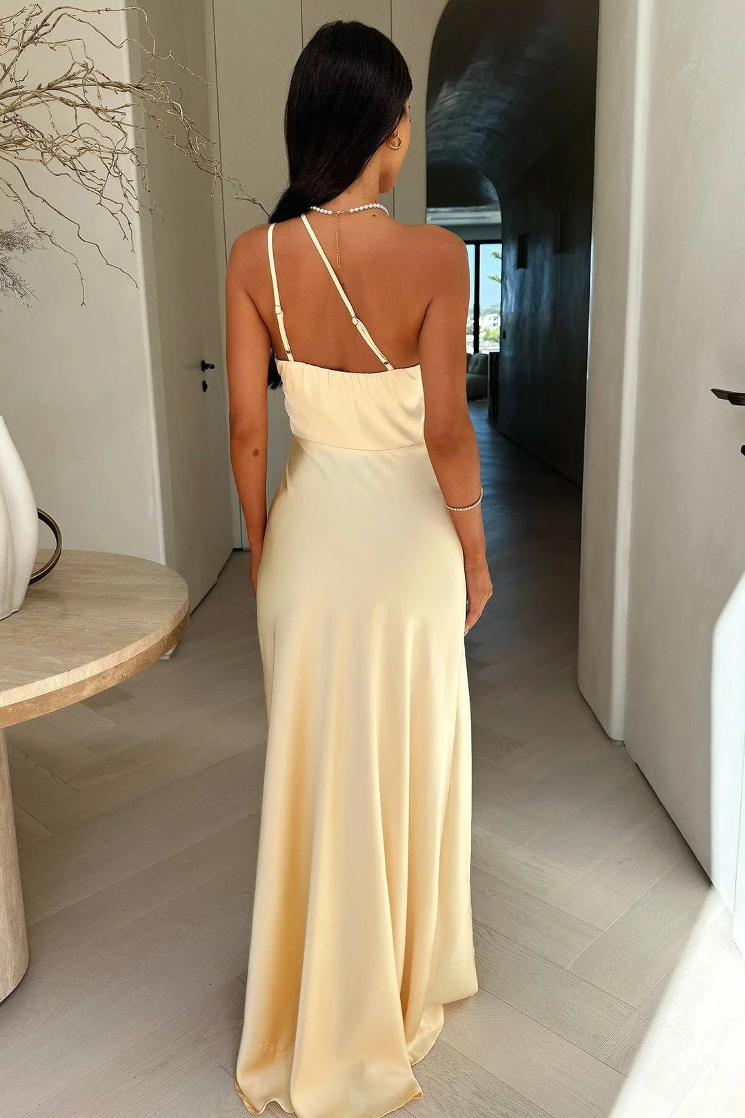 Back view of a cream satin one-shoulder maxi dress with delicate spaghetti straps and a fluid, floor-length silhouette.