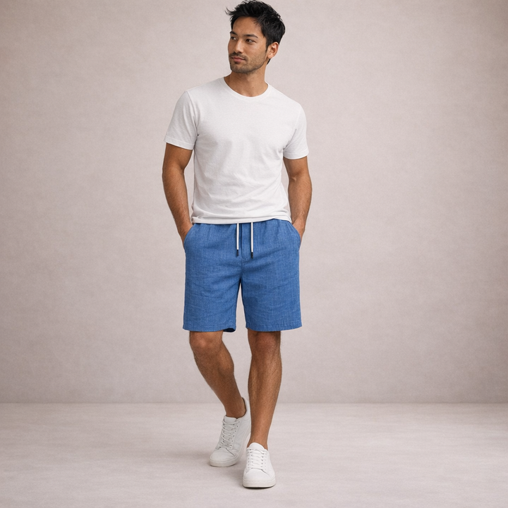 Man wearing blue linen-look drawstring shorts with a white t-shirt and sneakers against a neutral background.