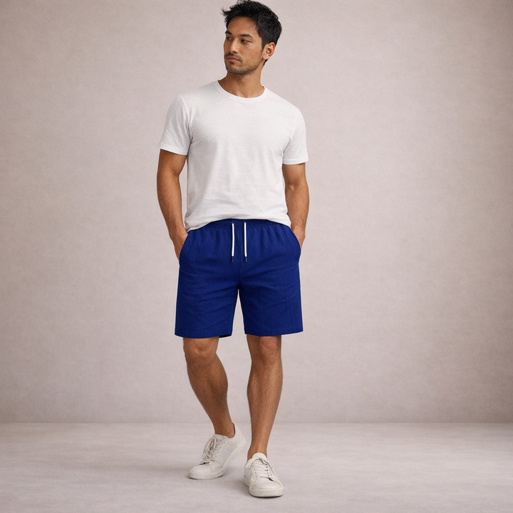 Man wearing blue linen-look drawstring shorts with a white t-shirt and sneakers against a neutral background.