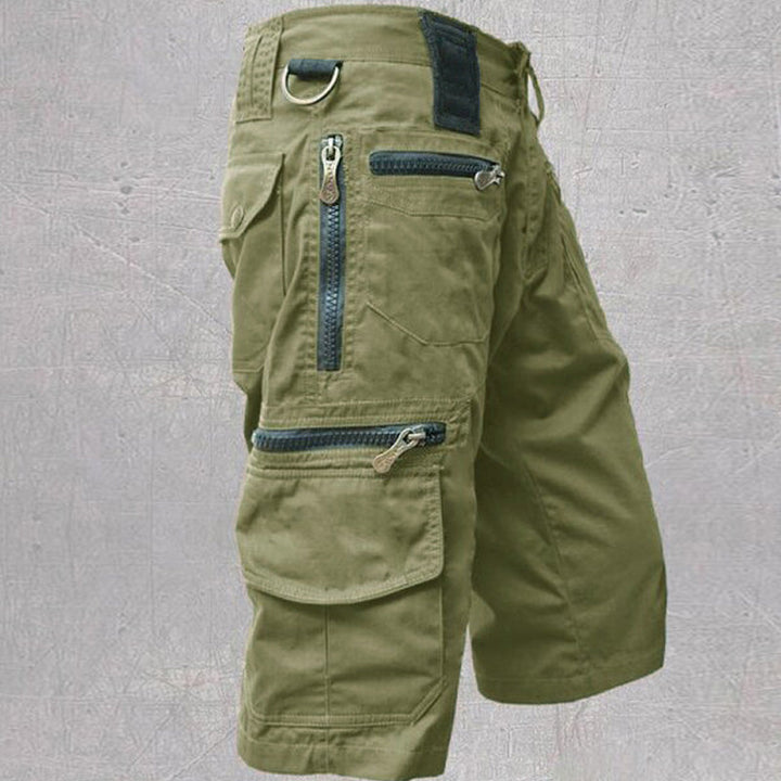 Olive green cotton cargo shorts featuring multiple zip and flap pockets, a D-ring, and a structured, durable utility design.