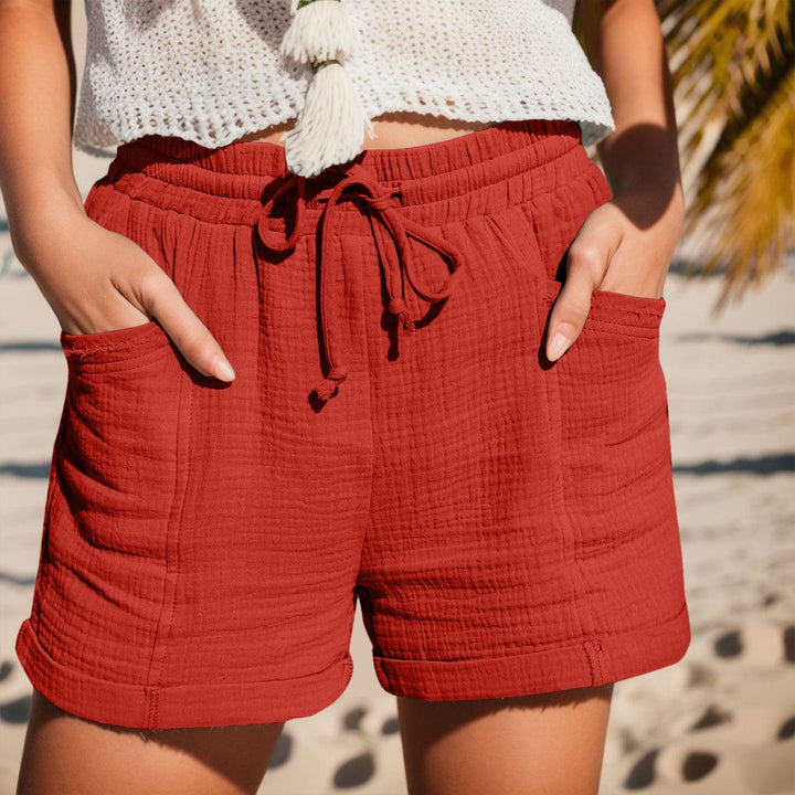 Rust-colored textured cotton shorts with an elastic drawstring waist, side pockets, and cuffed hems on a beach.