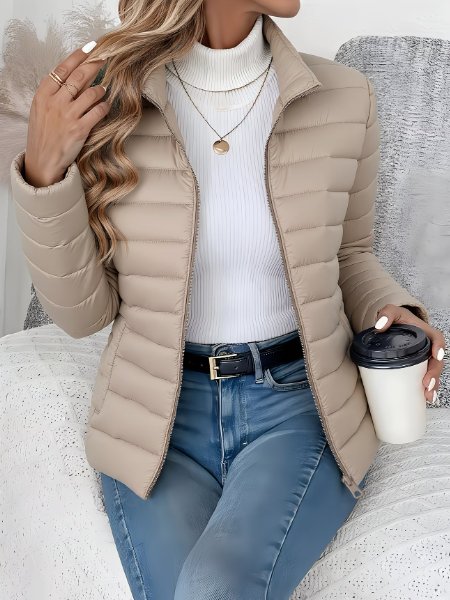 Beige quilted puffer jacket with a high stand collar, full-length zip, and tailored fit worn over a white turtleneck.