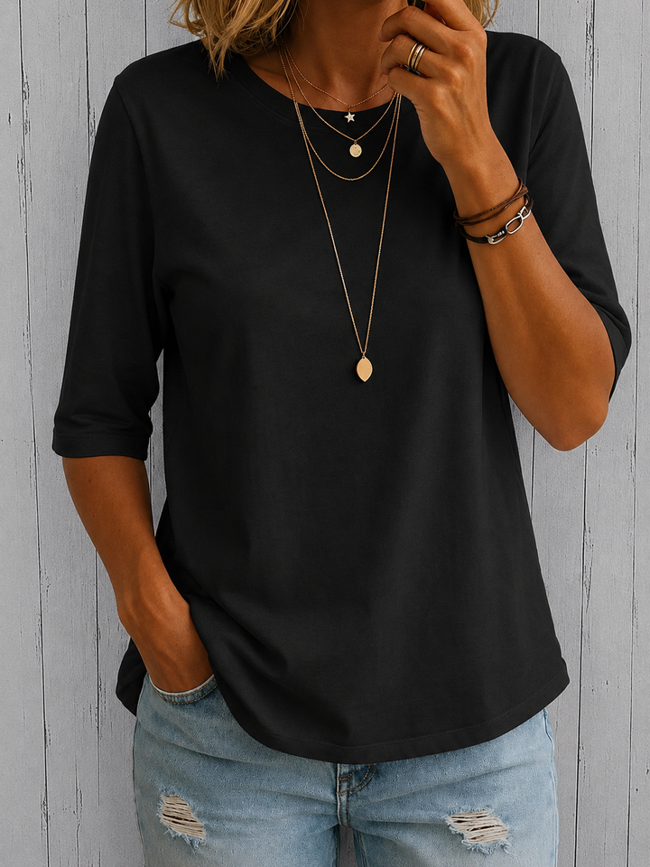 Woman wearing a black round-neck half-sleeve top with a relaxed fit and curved hem, styled with layered gold necklaces.