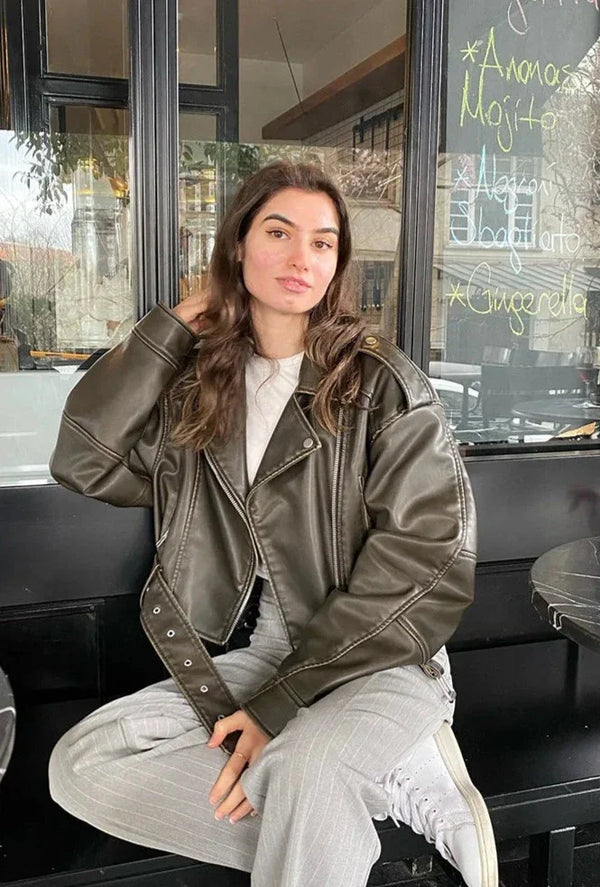 Woman wearing a cropped olive green faux leather biker jacket with an asymmetric zip and buckled waist belt.