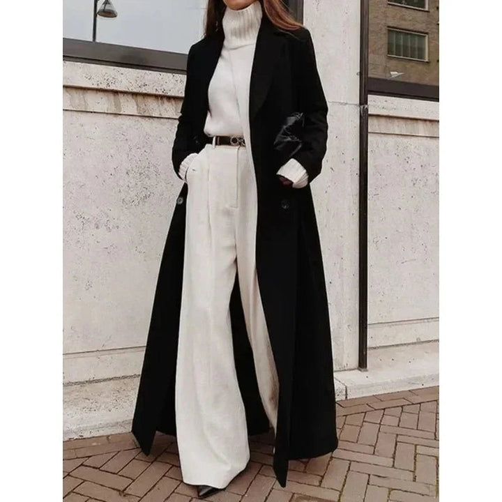 Woman wearing a black longline double-breasted coat with wide lapels over a white turtleneck and wide-leg trousers.