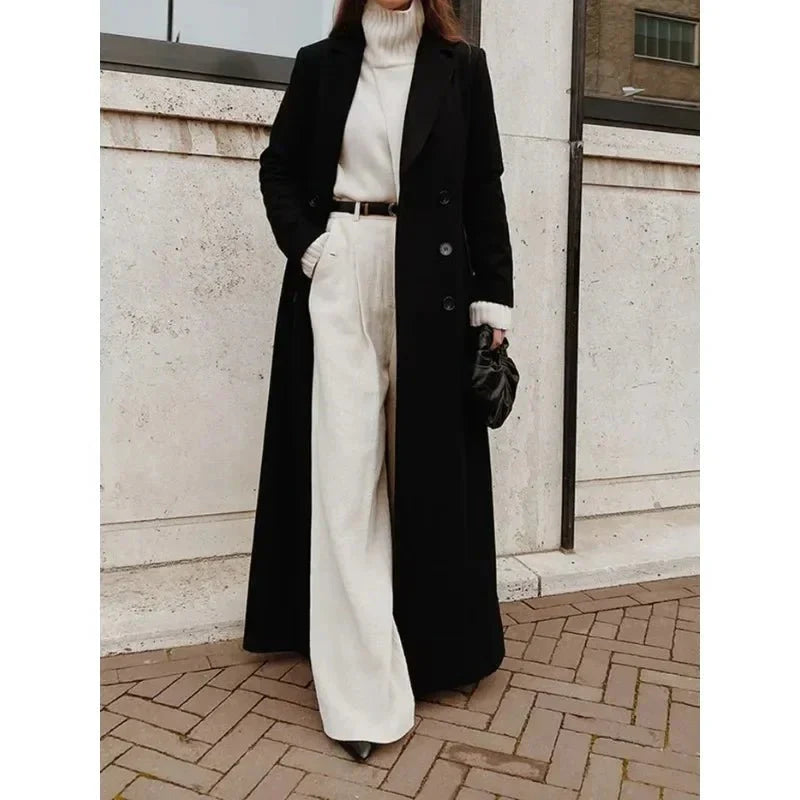 Black double-breasted longline coat with wide lapels worn over a white turtleneck and wide-leg trousers.