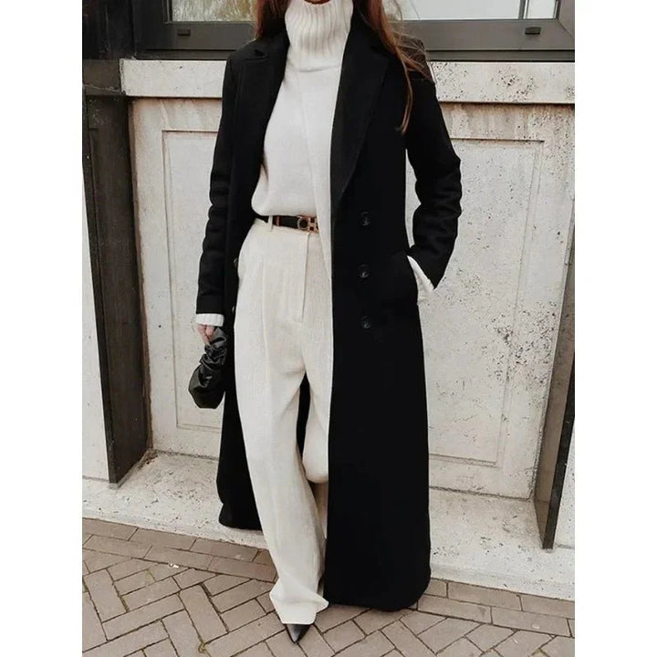Black longline double-breasted coat with wide notch lapels worn over a white turtleneck and tailored trousers.
