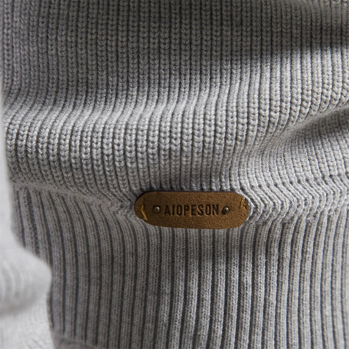 Close-up of a light grey ribbed knit sweater featuring a brown leather brand tag stitched to the hem.