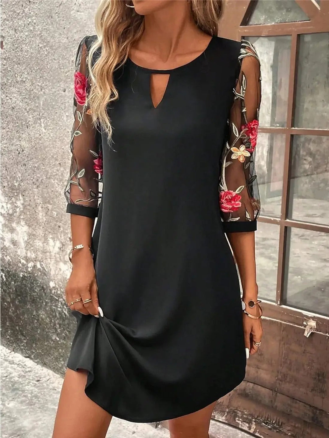Black mini shift dress featuring sheer mesh sleeves with colorful floral embroidery and a keyhole neckline.