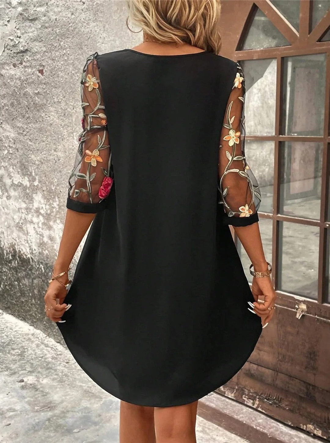 Back view of a black mini shift dress featuring sheer mesh sleeves with colorful floral embroidery and contrast cuffs.