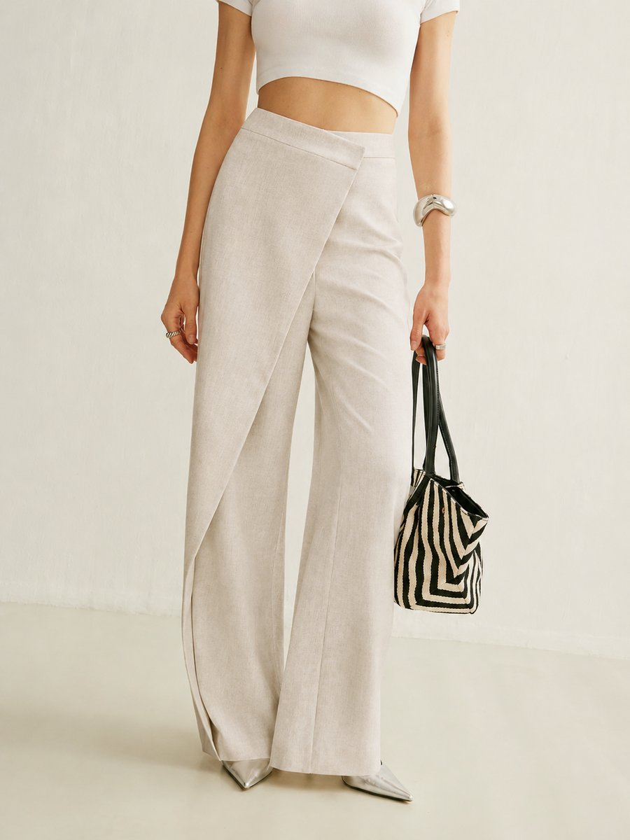Beige high-waisted wide-leg trousers with an asymmetric wrap front panel, styled with a white crop top and metallic heels.