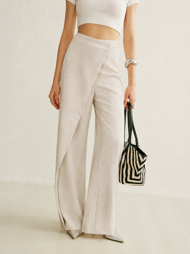 Beige high-waisted wide-leg trousers with an asymmetric wrap front panel, styled with a white crop top and metallic heels.