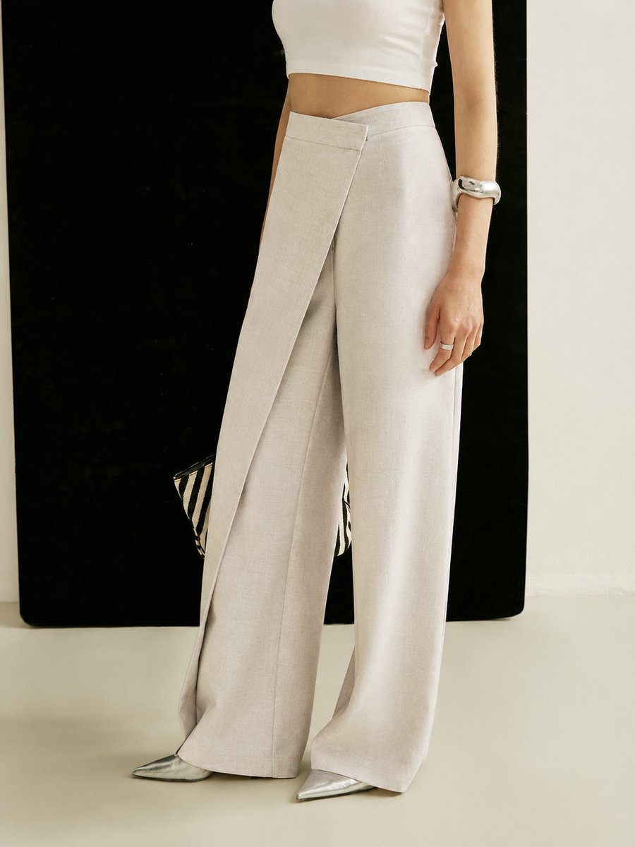 Beige high-waisted wide-leg trousers featuring a modern asymmetric wrap front panel in a smooth, tailored fabric.