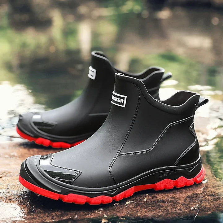 Black waterproof ankle rain boots with a rugged red treaded sole and rear pull tab, set on a wet outdoor surface.