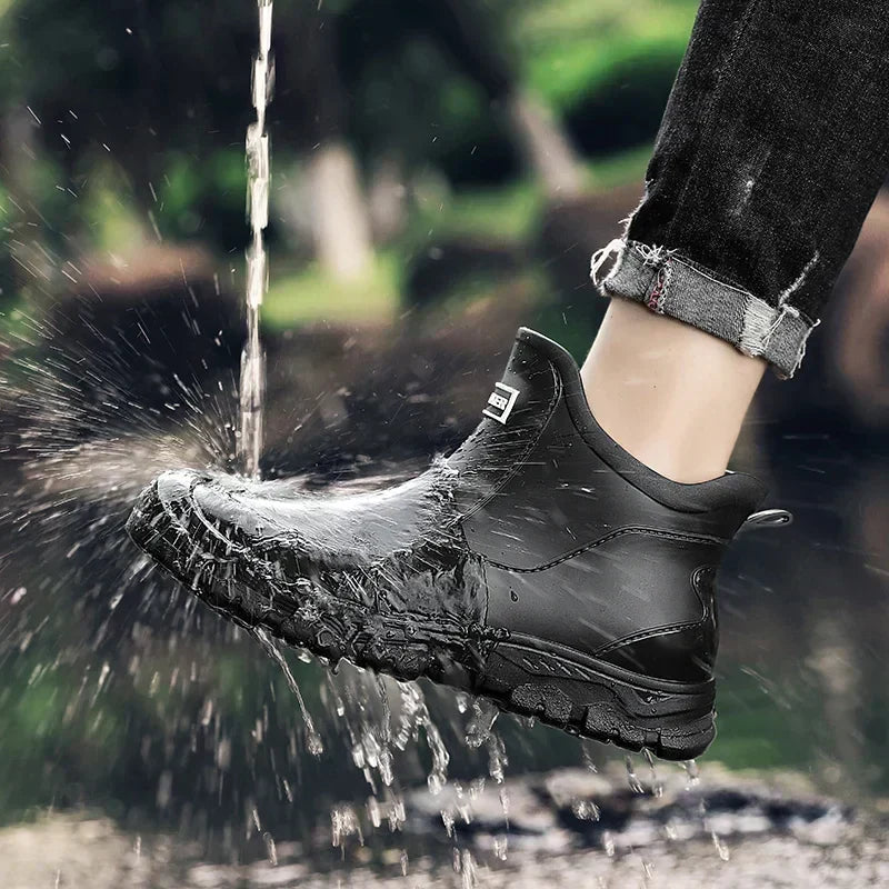 Black waterproof ankle rain boots with a rugged tread sole and pull tab, shown being splashed with water.