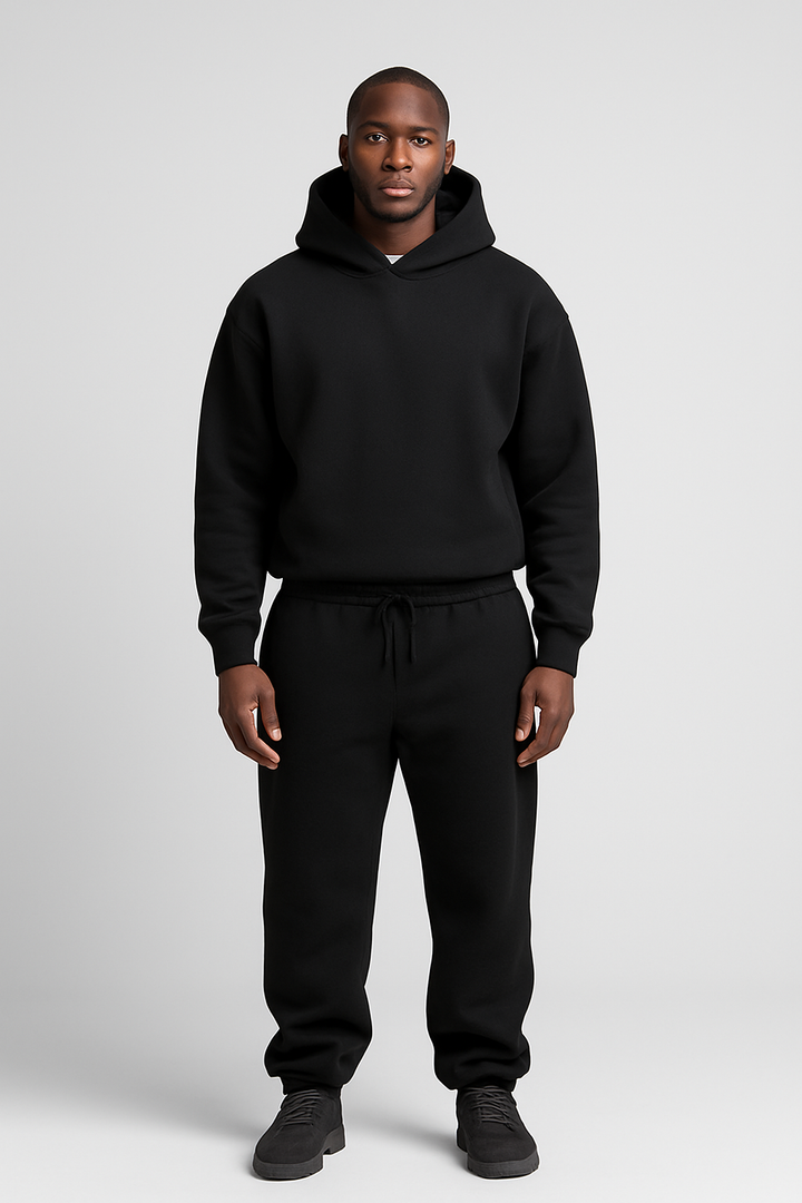 Man wearing a black cotton hoodie and matching drawstring joggers set against a plain light grey background.