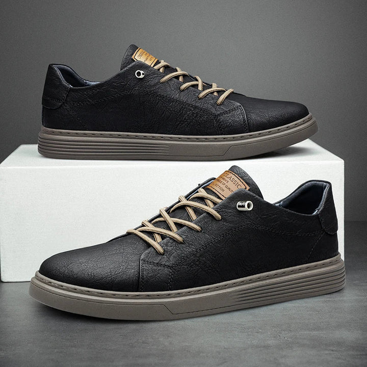 Black textured leather low-top trainers with beige laces and a ribbed grey sole, featuring a tan tongue label.