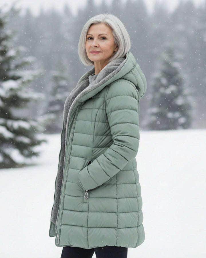 Woman wearing a sage green longline puffer coat with a cozy fleece-lined hood in a snowy outdoor setting.