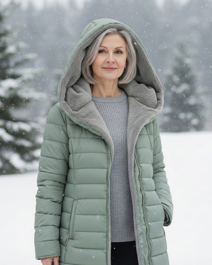 Woman wearing a sage green longline puffer coat with a cozy grey fleece-lined hood in a snowy outdoor setting.