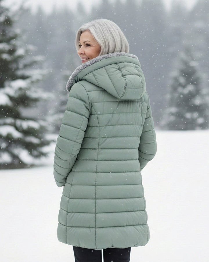 Woman wearing a sage green longline puffer coat with a cozy fleece-lined hood in a snowy outdoor setting.