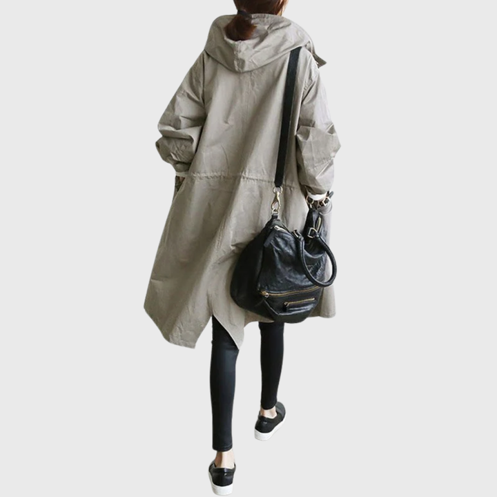 Woman wearing an oversized beige hooded waterproof raincoat with a longline silhouette and black shoulder bag.