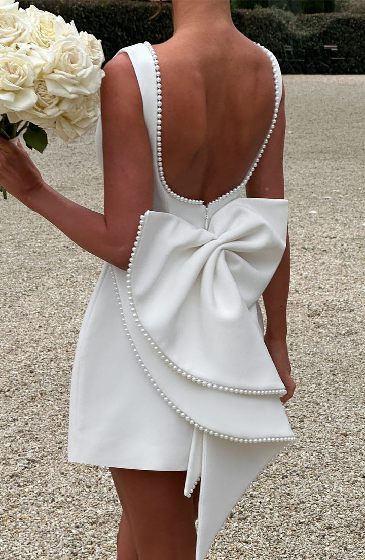 White sleeveless mini dress with pearl-trimmed back, scoop neckline, and a large statement bow detail.