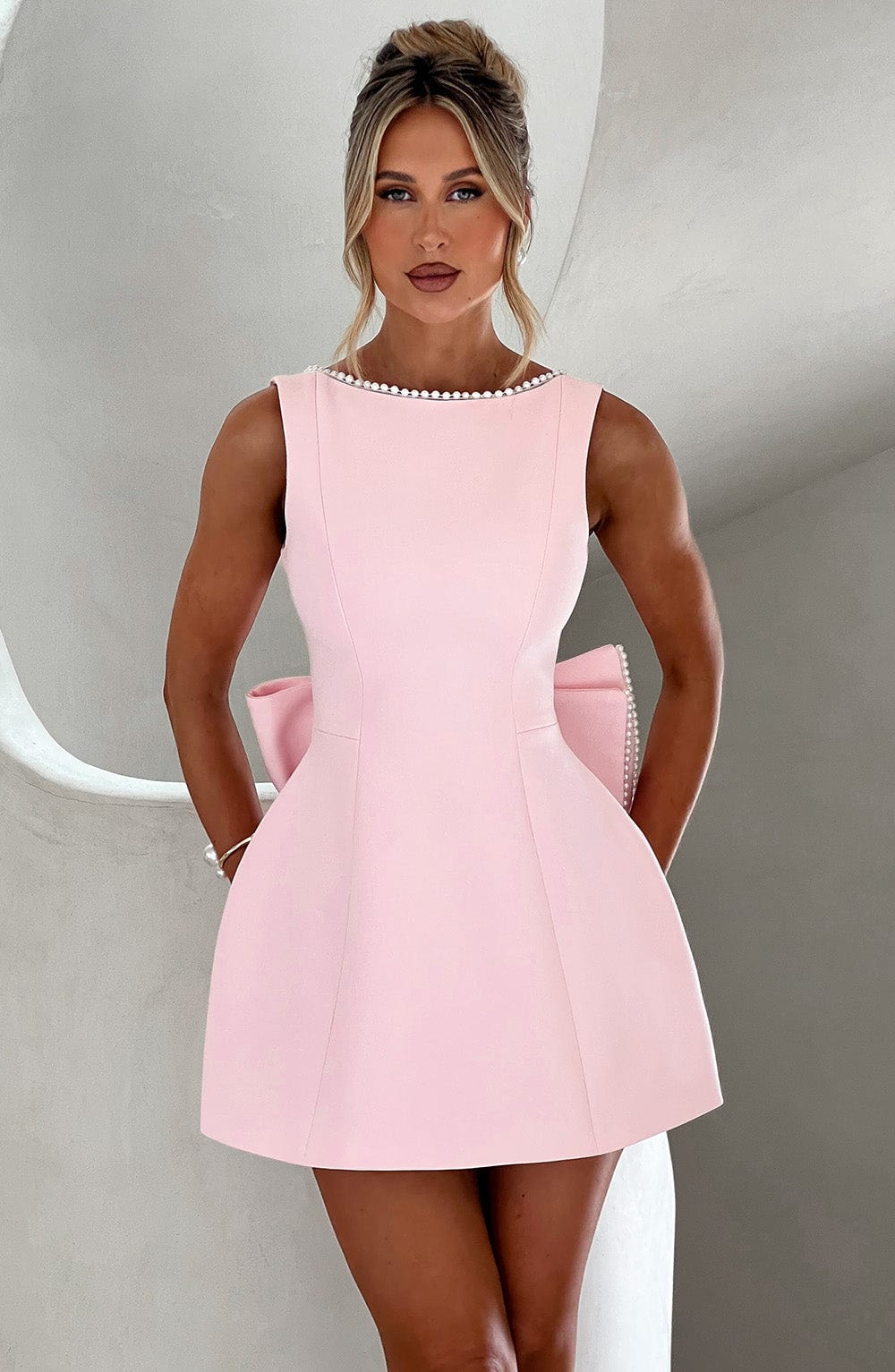 Sleeveless light pink mini dress featuring a pearl-trimmed neckline and a large structural bow detail at the back.