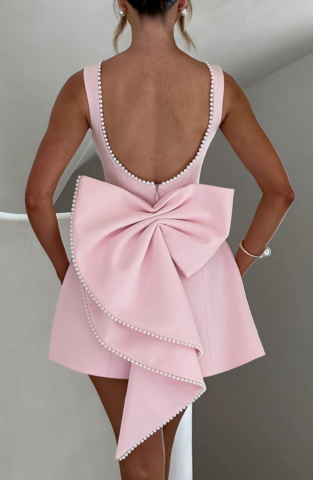 Back view of a sleeveless pink mini dress with pearl trim, a low scoop back, and a large statement bow detail.