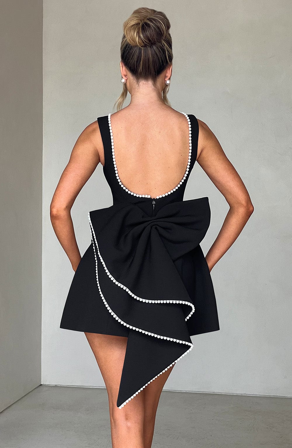 Back view of a black sleeveless mini dress with pearl trim, a low scoop back, and a large statement bow detail.