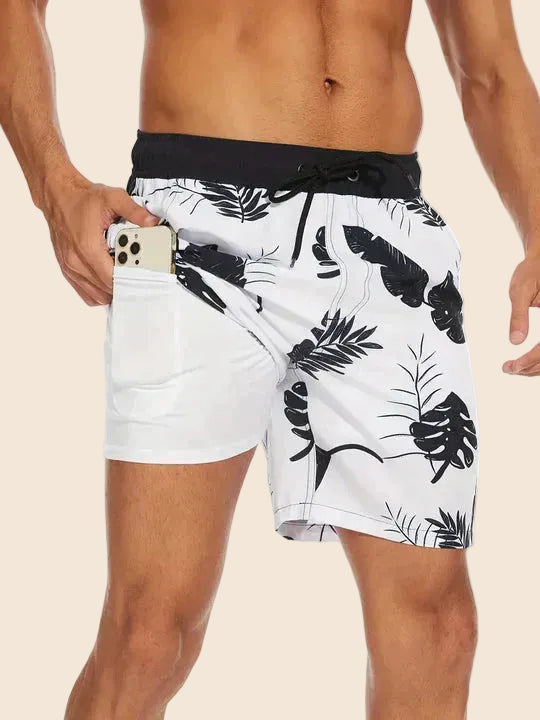 Men's white swim shorts with black leaf print, drawstring waistband, and a built-in liner with a side phone pocket.