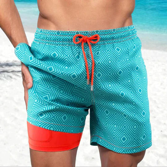 Teal geometric print swim shorts with a bright orange drawstring and matching inner liner on a beach background.