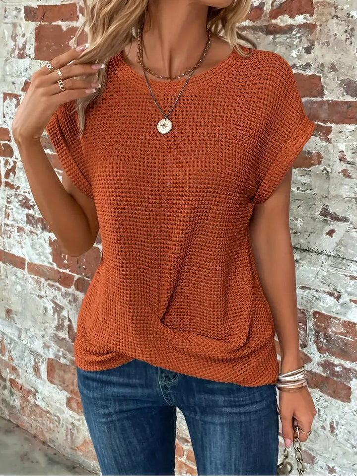 Rust orange waffle knit top with rolled cap sleeves and a flattering twist-front hem, styled with blue jeans.