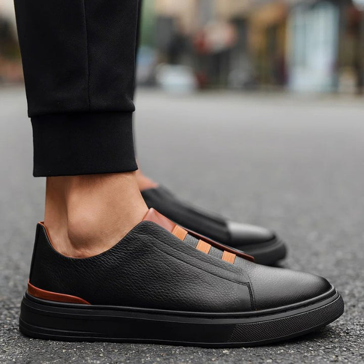Men's black leather slip-on trainers featuring brown elastic strap detailing and a durable black rubber outsole.