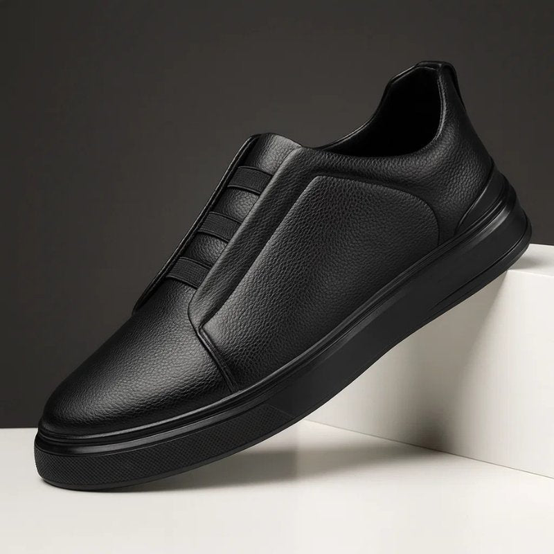 Black leather slip-on trainers with a textured finish, elastic strap detailing, and a durable rubber sole.