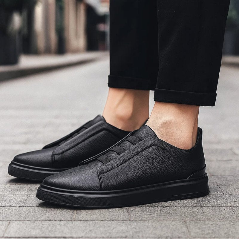 Men's black leather slip-on trainers with elastic straps and a durable rubber sole, worn with black trousers.