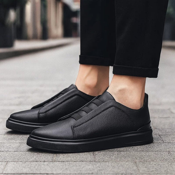 Men's black leather slip-on trainers with elastic straps and a durable rubber sole, worn with black trousers.
