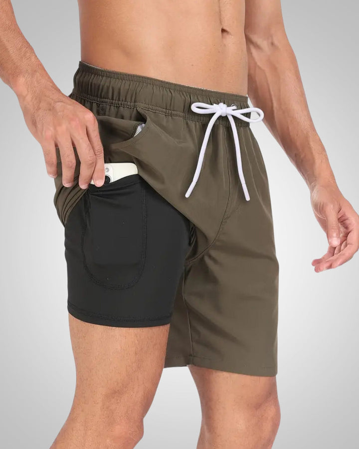 Olive green swim shorts with a white drawstring and black built-in compression liner featuring a side phone pocket.