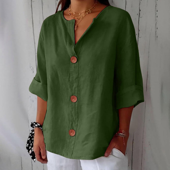 Olive green linen V-neck blouse with wooden buttons and relaxed three-quarter sleeves worn with white trousers.